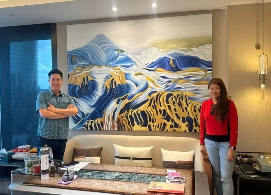 Two women standing in a modern living room with a large mountain landscape painting. The woman on the left is wearing a blue patterned shirt, and the woman on the right is wearing a red top and jeans. A beige sofa with assorted pillows is in front of the painting, and there are various items on the table in front of them.