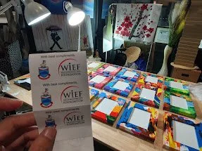 Two business cards held in front of a table filled with colorful craft or artwork pieces, with a window and areas of light in the background.