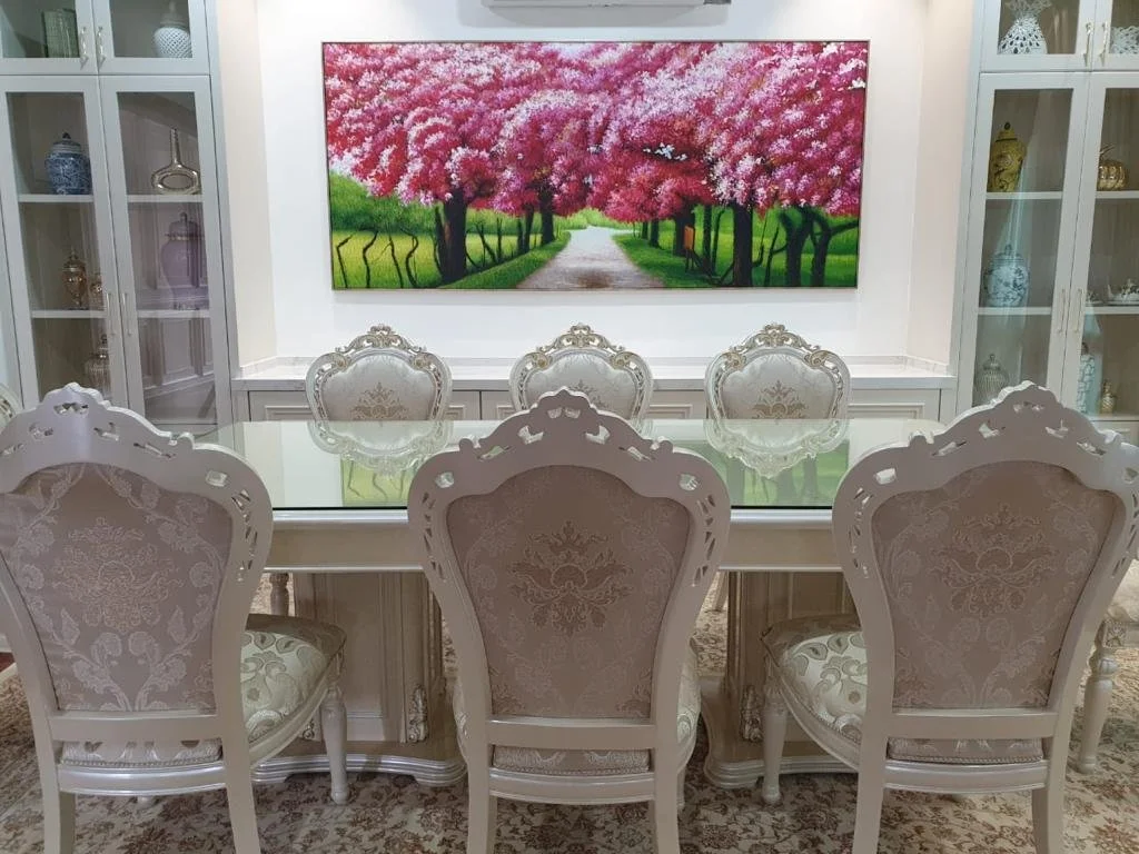 A dining room with six ornate white chairs with beige upholstery around a glass-top dining table. The room has glass-front cabinets with decorative vases and ceramics. A large painting of a pink blossoming tree-lined pathway hangs on the wall.