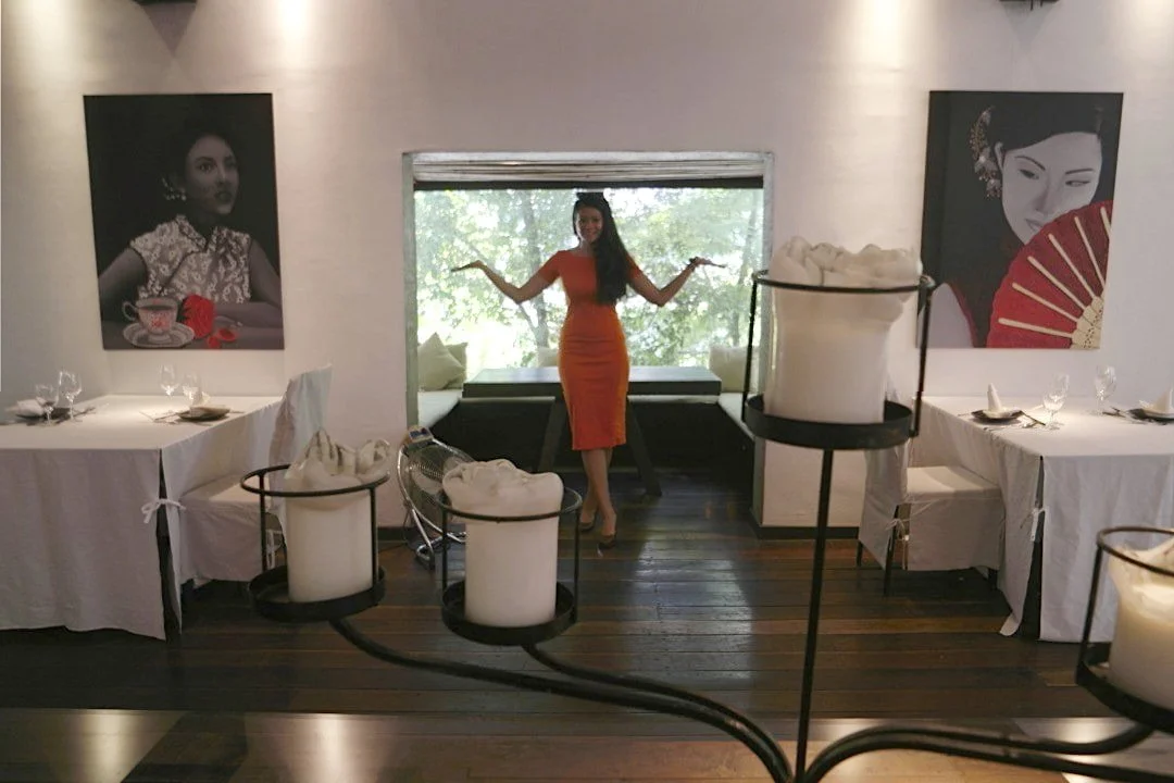 A woman in an orange dress standing inside a restaurant, balancing with her arms out, with two portraits of women on the wall behind her and a window showing greenery outside.