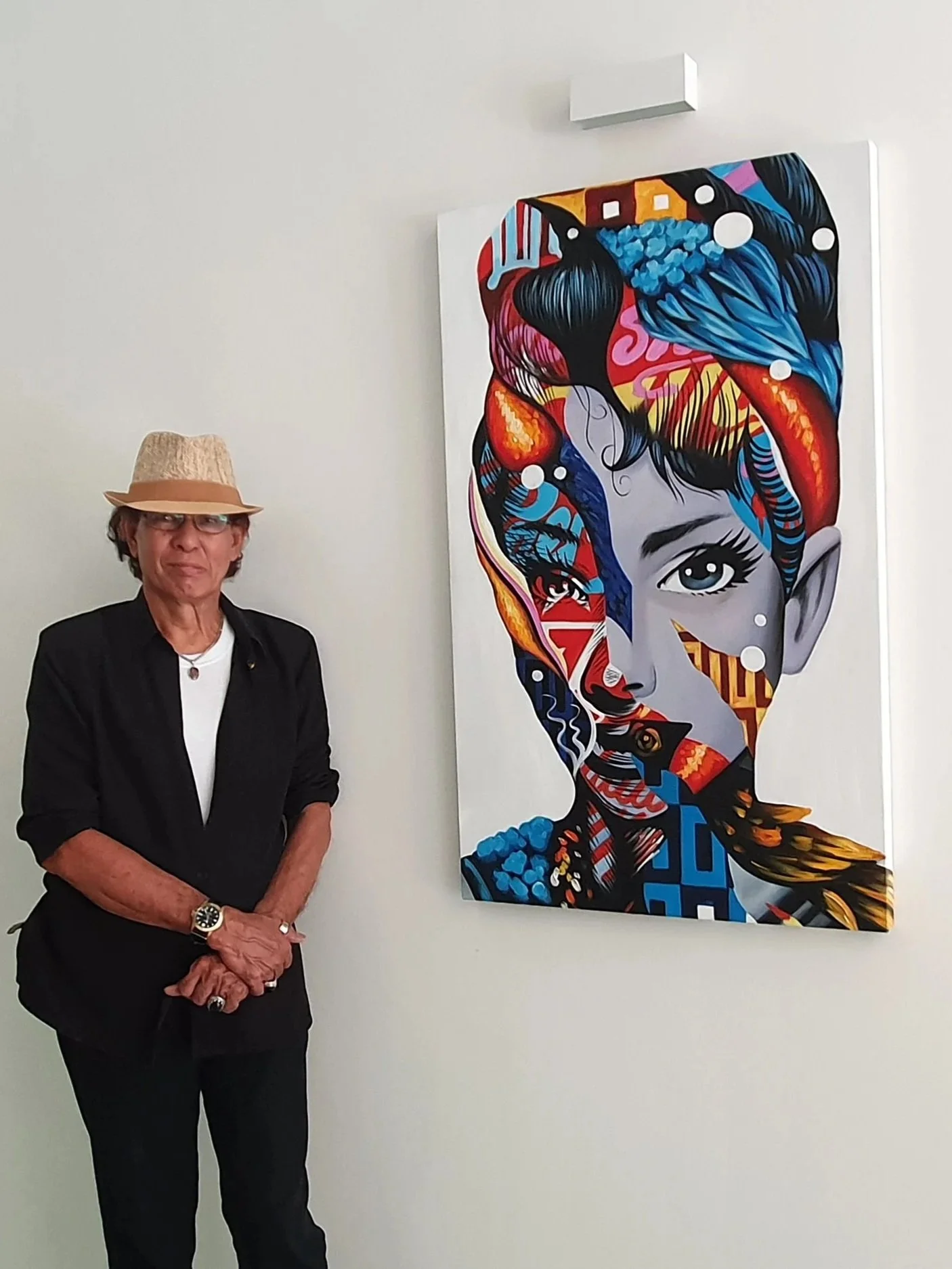 A woman wearing a beige hat, glasses, and a black blazer stands beside a colorful abstract portrait of a woman's face on a white wall.