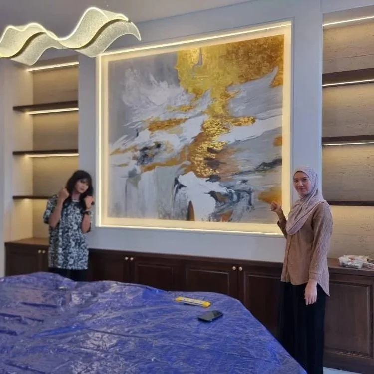 Two women standing in front of a large abstract mural with gold, white, black, and gray colors in a modern room with wood accents and lighting. One woman is wearing a hijab and smiling, the other is holding her hair, dressed in patterned clothes.