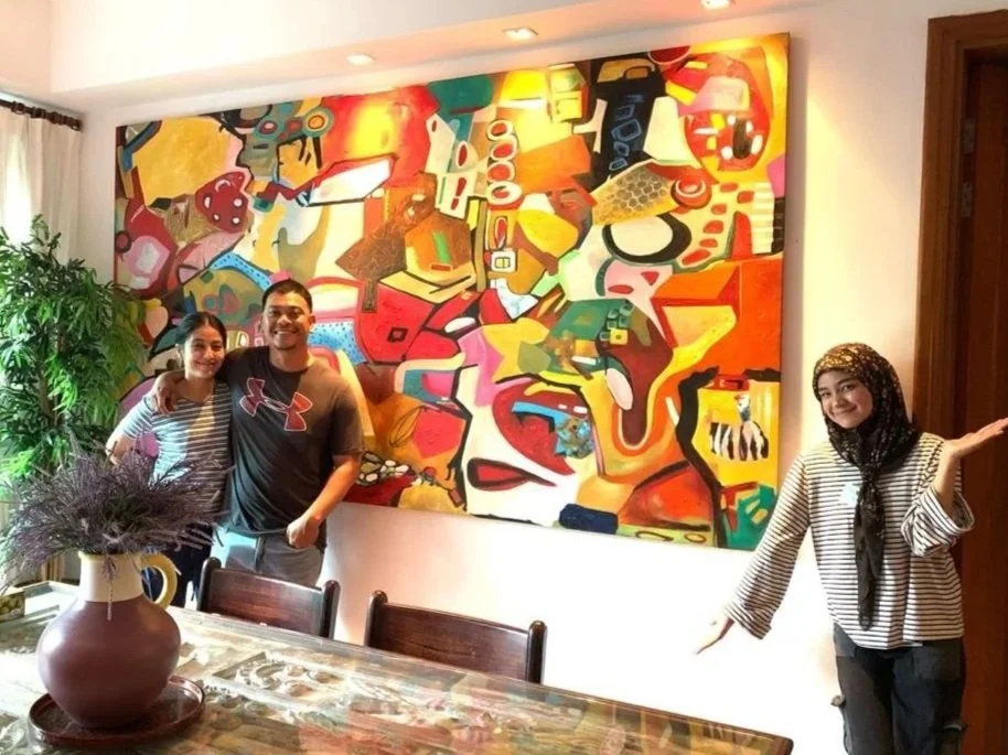 Three people standing in front of an abstract colorful painting in a well-lit room with a large vase of flowers on the table in foreground.