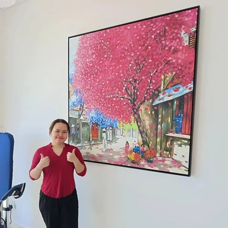 A woman giving a thumbs up while standing next to a large colorful painting of a street scene with pink, blue, and red trees and small houses.