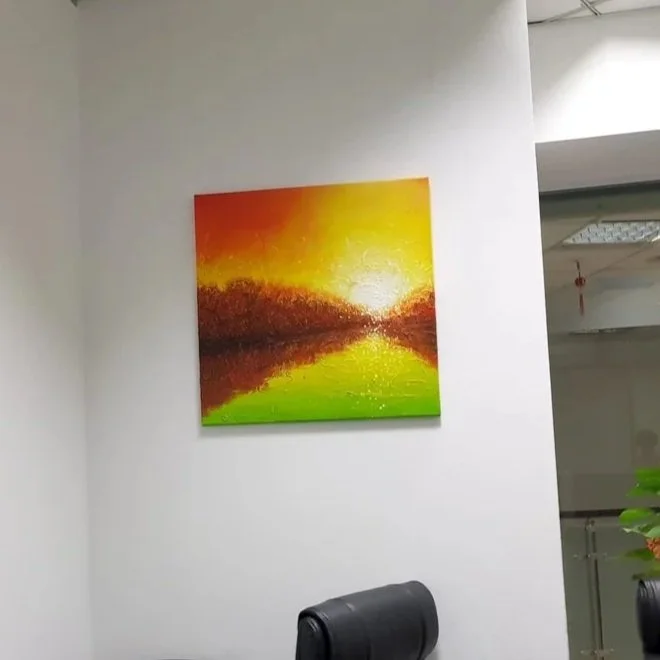 Colorful landscape painting featuring a sunset over a lake with green, orange, and yellow hues, hung on a white wall in an office setting.