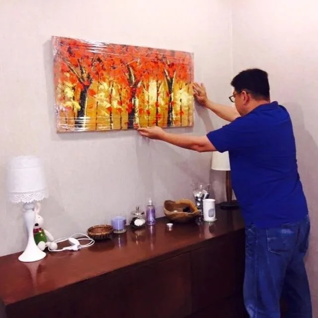 A man in a blue shirt and glasses is removing plastic wrap from a framed painting of an autumn forest scene on a white wall.