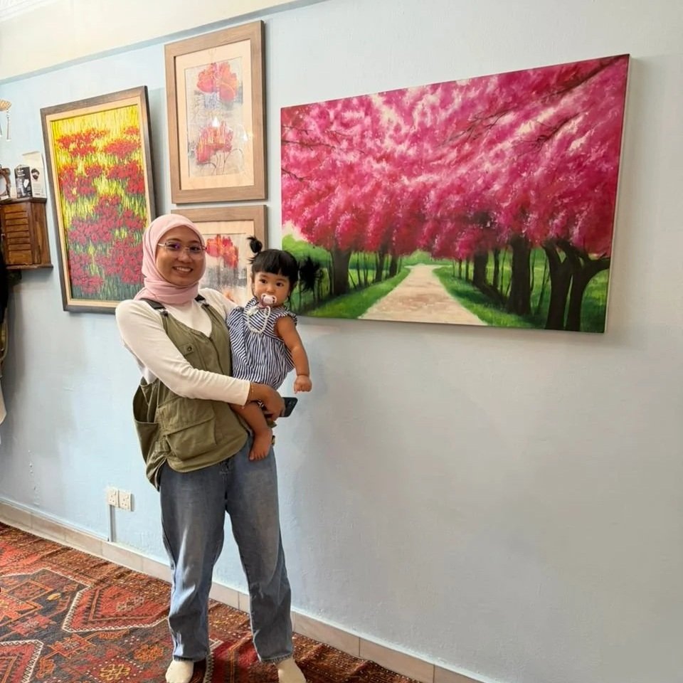 A woman wearing a pink hijab and glasses holding a young girl, both smiling, standing in front of a mural of a tree-lined path with pink blossoms on the wall of an art gallery.