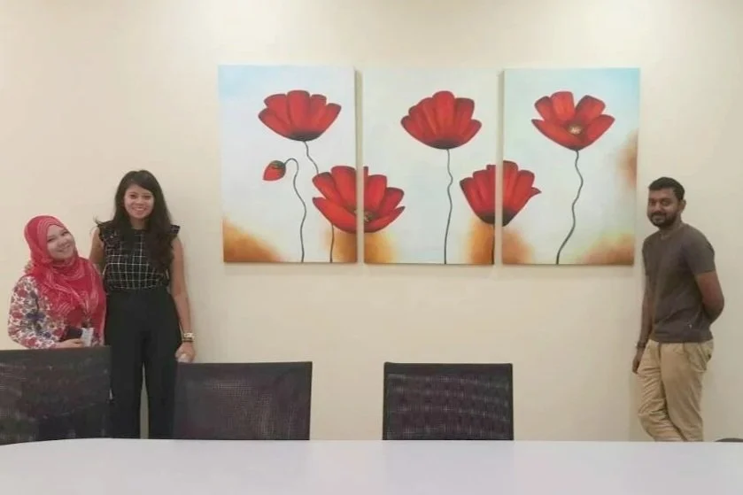 Three people standing in a room with a large painting of red flowers on the wall behind them, two women and one man, with chairs and a table in the foreground.