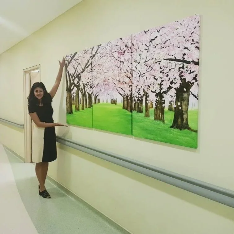 A woman in a black dress stands beside a three-panel painting of cherry blossom trees on a green field, gesturing towards it with a smile.