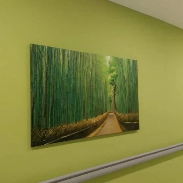 A painting of a forest with a dirt path, green trees, and sunlight, hanging on a light green wall.