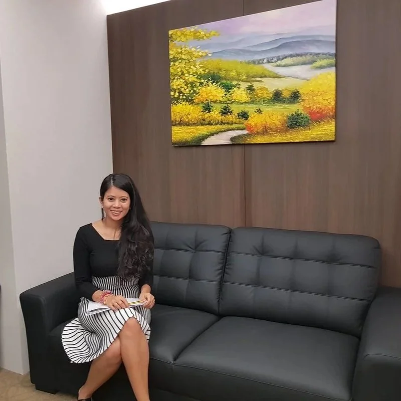 A woman sitting on a black sofa in a room with wooden paneling and a colorful landscape painting on the wall behind her.