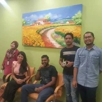Four people in a room with green walls, two women and two men, sitting and standing around a large painting of a sunny rural landscape with a flower field and a winding path.