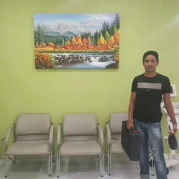 A man standing next to three beige waiting room chairs against a lime green wall with a colorful landscape painting of mountains, trees, and a river.
