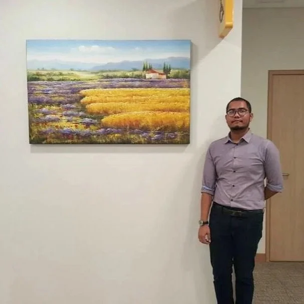 A man in a light purple button-up shirt and dark pants standing beside a wall with a large landscape painting of fields, trees, and mountains.