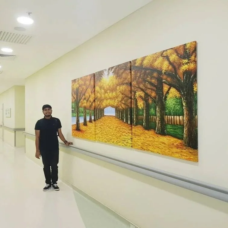 A man standing next to a multisection painting of a tree-lined pathway during autumn.