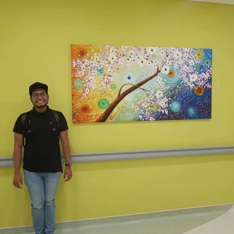 A man standing next to a colorful abstract painting of a tree with blooming pink flowers on a yellow wall.
