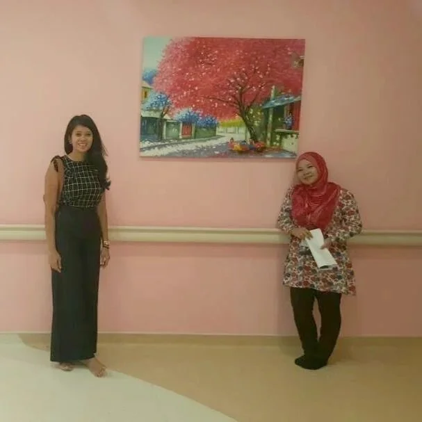 Two women standing in front of a pink wall with a colorful painting of a tree with pink leaves.
