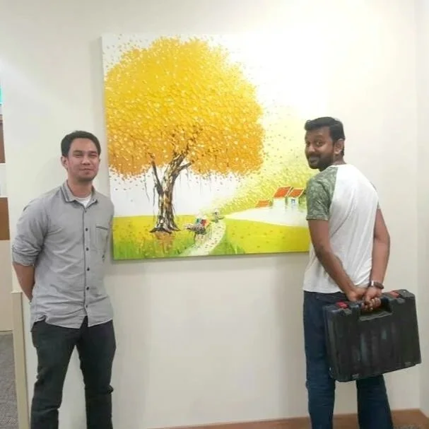 Two men standing in front of a painting of a yellow-leaved tree, a small path, and red-roofed houses in a rural landscape.