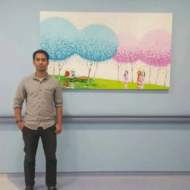 Young man standing in front of colorful artwork depicting trees and a pond with two women standing nearby.