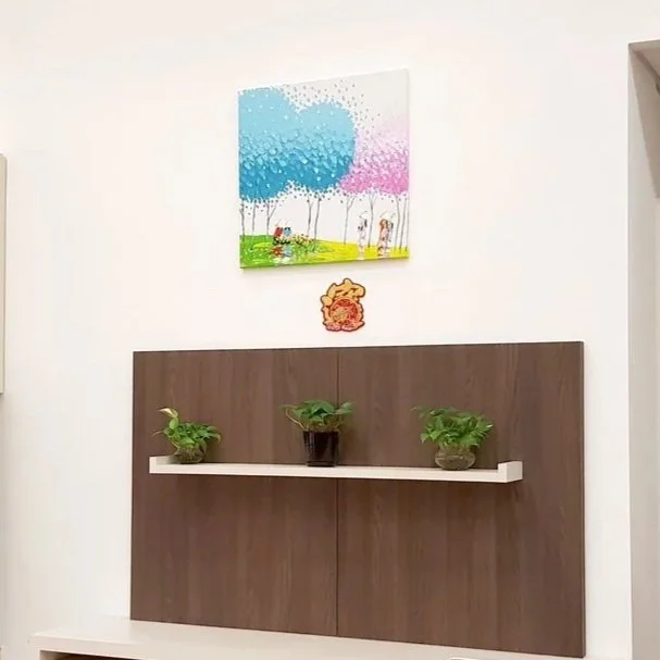 Three potted plants on a white shelf mounted on a wooden panel on a white wall. Above the shelf is a colorful abstract painting and a small decorative wall hanging.