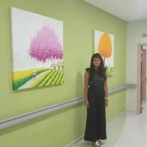 A woman standing in a hallway with bright green walls, next to two paintings of trees with pink and orange foliage, respectively.