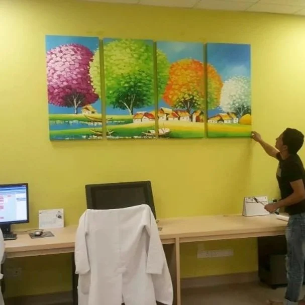 An office with yellow walls, a long desk with a white coat hanging over a chair, a desktop computer, and a person pointing at a five-panel landscape painting of colorful trees and a small village.