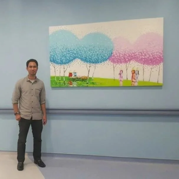 A man standing next to a colorful landscape painting on a light blue wall.