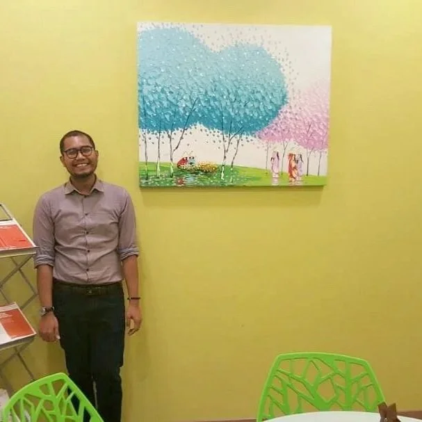 A smiling man in glasses standing next to a colorful painting of trees and children on a yellow wall, with green chairs nearby.