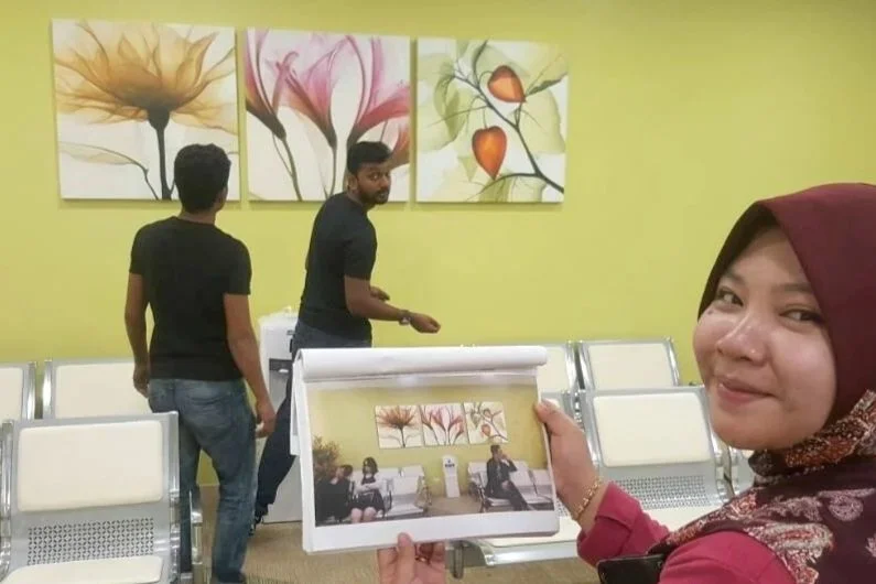 A woman wearing a maroon hijab and red shirt smiling and holding a photo of herself with two men, inside a waiting room with yellow walls and floral artwork on the wall.