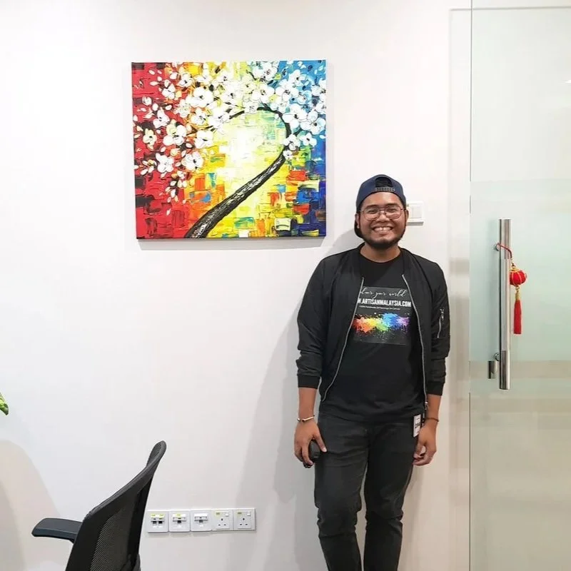 A smiling man wearing glasses, a black jacket, and a baseball cap standing in an office next to a colorful abstract painting of a cherry blossom tree on a white wall.