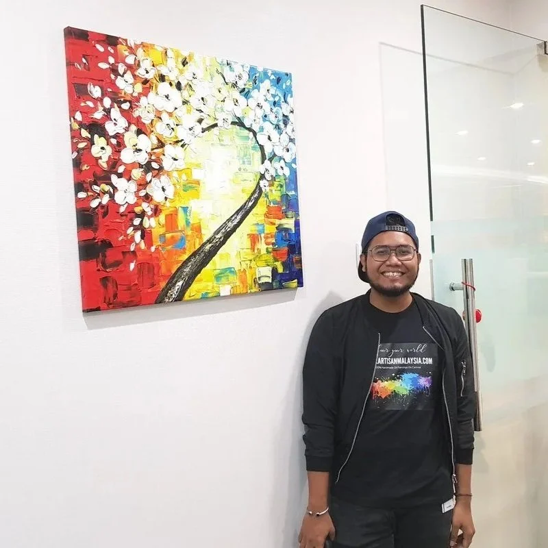 A young man with glasses and a cap smiling, standing next to a colorful abstract painting of a blossoming tree on a white wall.