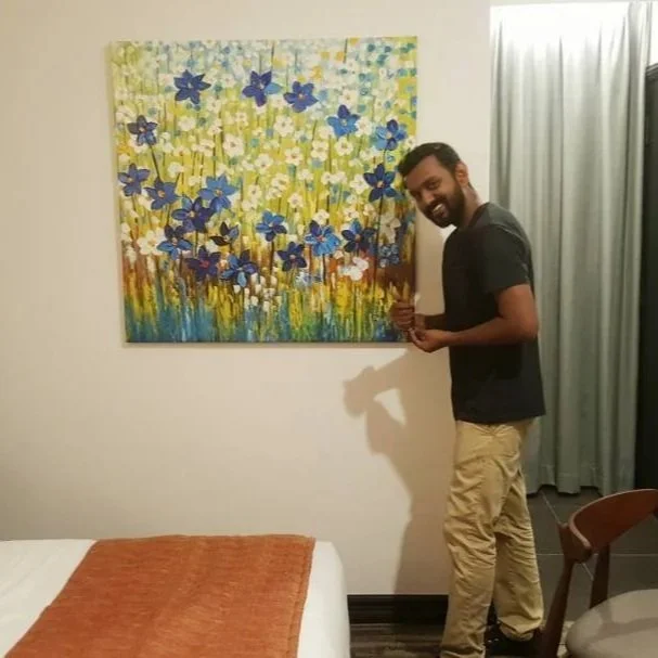 A man standing next to a colorful floral painting on a wall, smiling and holding a drink in a room with beige walls, a bed with an orange blanket, and a curtain.