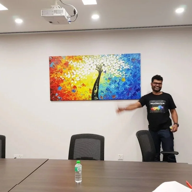 A man standing in a conference room, pointing at a colorful abstract painting of a tree with white, orange, yellow, green, blue, and red leaves on a white wall.