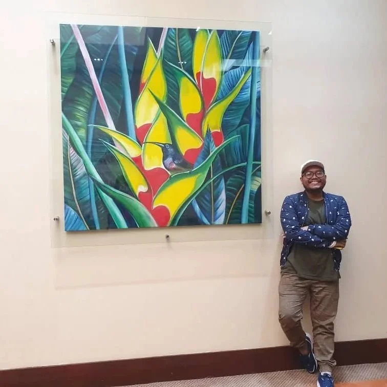 A man standing with arms crossed, smiling, next to a colorful painting of a toucan bird nestled among large tropical leaves.