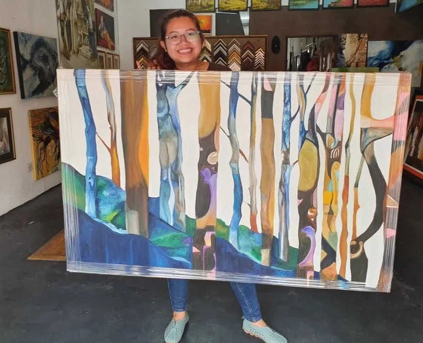 A woman holding a colorful abstract painting of a forest scene with tall, slender trees in a gallery with other artwork on the walls.