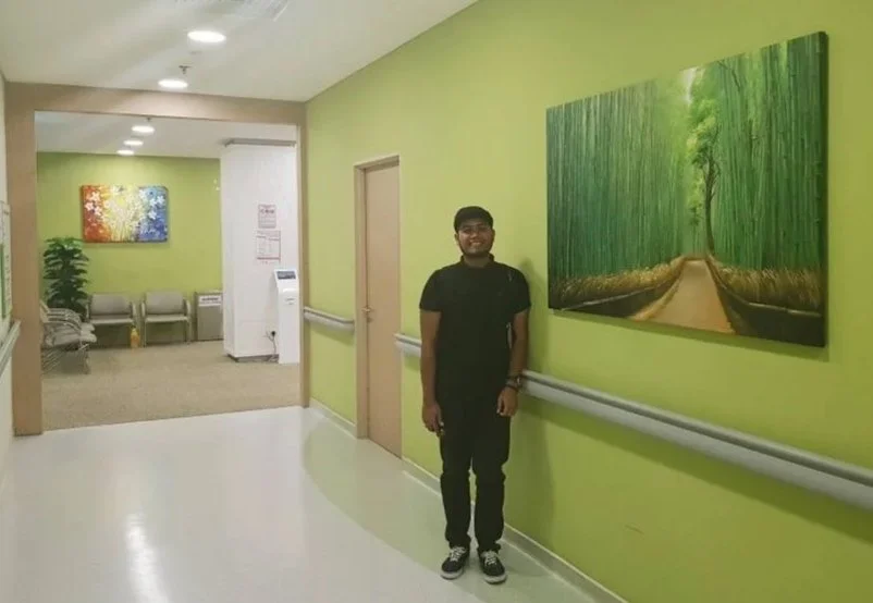 A young man standing in a hospital corridor with green walls, smiling. There is a large framed photograph of a forest pathway on the wall behind him, and in the background, there is artwork, seating area, and an air purifier.