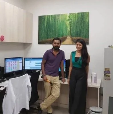 Two people, a man and a woman, standing in an office with computer monitors and a painting of a forest path hanging on the wall behind them.