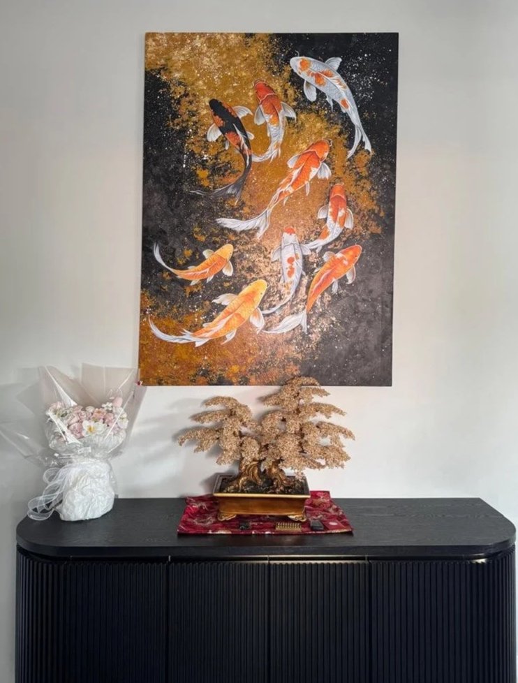 A black sideboard with a decorative gold-colored tree in a rectangular pot, a red cloth underneath, and a bouquet of pink and white flowers in a wrapped bouquet on the left. Above, a painting of colorful koi fish swimming against a black and gold bac