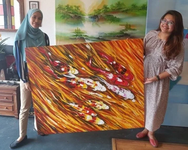 Two women hold a large abstract painting of colorful swirling red, orange, yellow, and white brushstrokes on a wooden background. One woman wears a light blue hijab and beige pants, the other wears glasses and a light-colored dress.