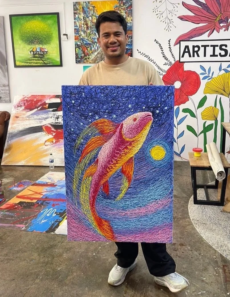 A young man holding a colorful painting of a koi fish under a starry sky, with a yellow moon. The background has vibrant blue, pink, and purple hues, and there are various artworks and paintings displayed on the walls around him.