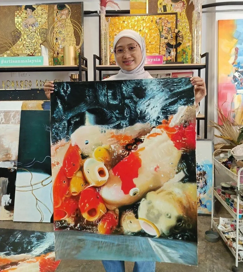 A woman wearing a white hijab and glasses is holding a colorful painting of koi fish swimming in water. She is smiling and standing in an art studio with various artworks and supplies in the background.