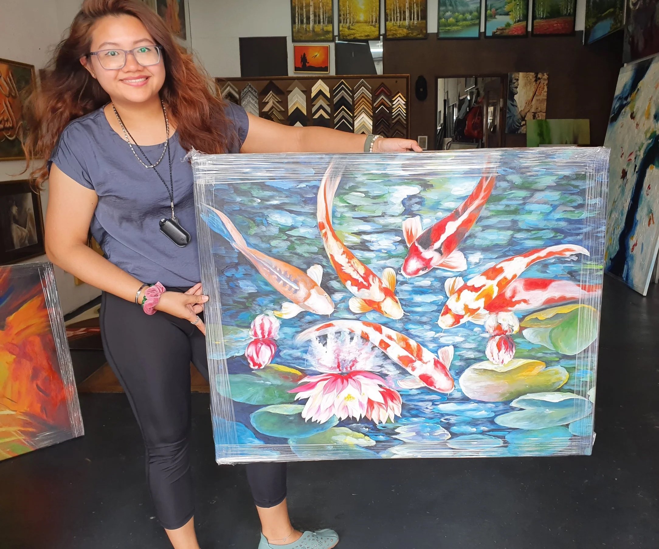 A young woman with glasses, long curly hair, and a gray T-shirt is standing in an art gallery, smiling and holding a colorful painting of koi fish swimming in water with lily pads.