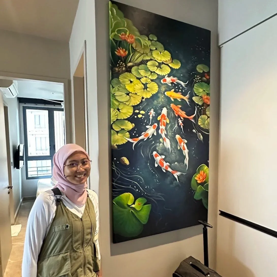 A woman with glasses and a pink headscarf standing next to a large painting of koi fish swimming among lily pads and flowers in water.