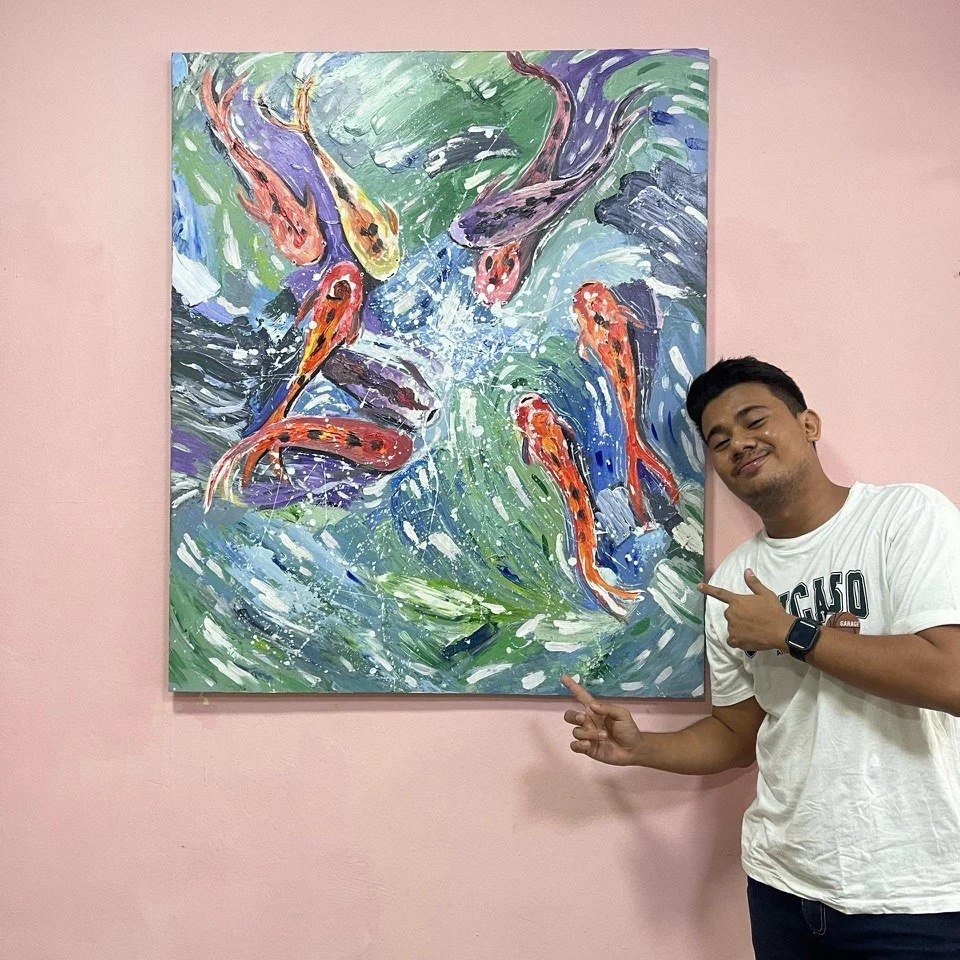 A young man in a white T-shirt pointing at a colorful painting of koi fish swimming in water, hung on a pink wall.