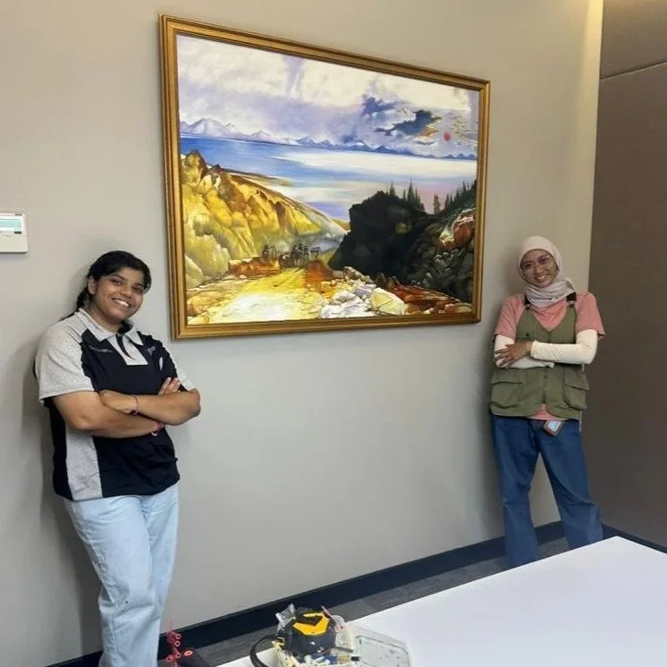 Two women stand on either side of a framed landscape painting in a room with gray walls, smiling with arms crossed, one in a striped shirt and the other in a pink shirt and hijab.