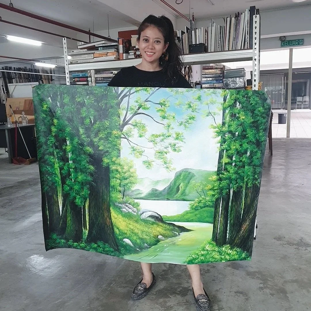 A woman holding a large landscape painting of a forest with trees, mountains, and a river.