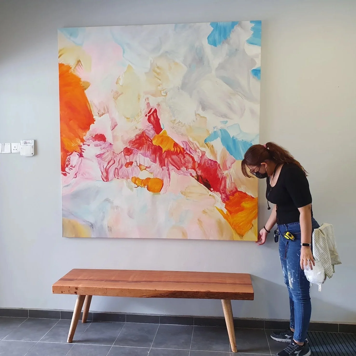 A woman wearing black shirt, jeans, and a face mask stands beside a large abstract painting on a wall. The painting features bright, vibrant swirls of red, pink, yellow, orange, blue, and white colors.