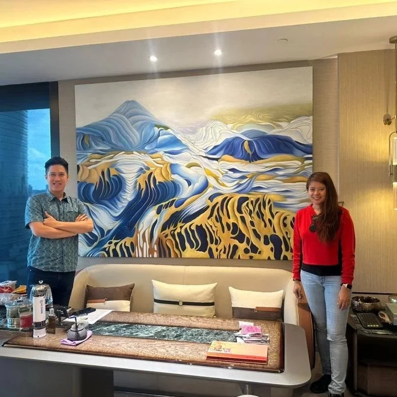 Two people standing in front of a mountain landscape painting in a room with a sofa, table, and various items on the table.