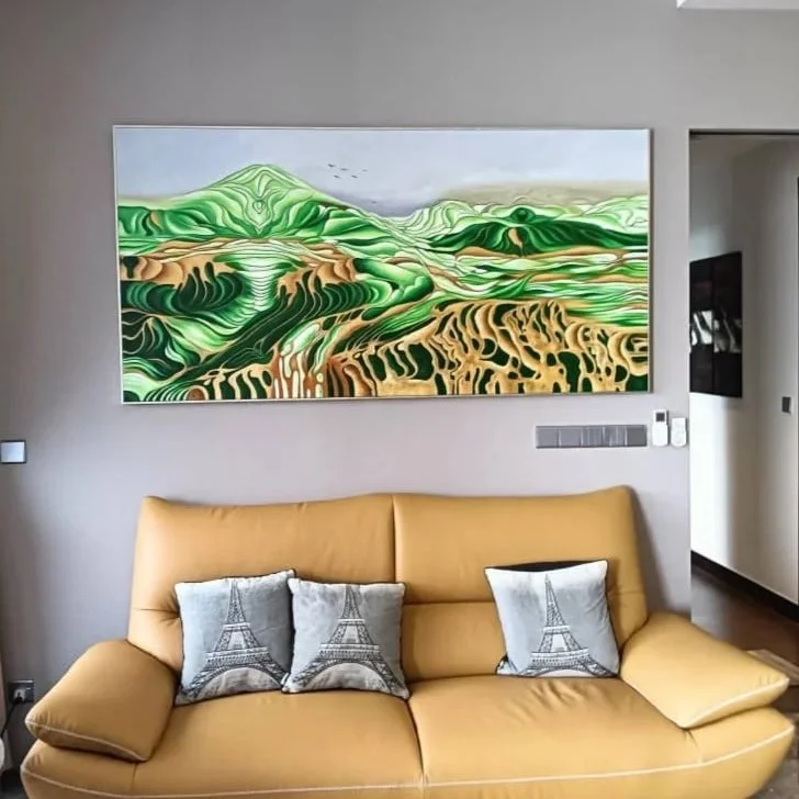 Modern living room with a large abstract landscape painting of green hills and fields above a tan sofa. The sofa has three cushions with Eiffel Tower designs. The room has neutral walls and some wall switches.
