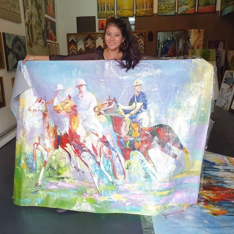 A woman smiling and holding up a colorful painting of three horse riders in a style reminiscent of impressionism, in an art gallery surrounded by framed paintings.